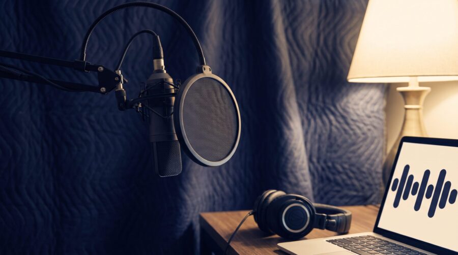 Home audiobook recording setup with a dynamic microphone, pop filter, closed-back headphones, and a laptop running audiobook recording software beside draped moving blankets